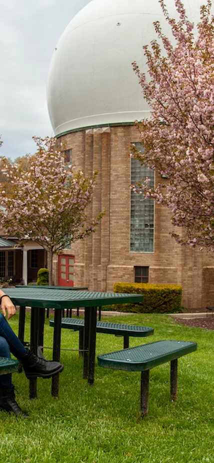 Kara Brugman sits at a picnic table on the Broad Branch Road campus, the home of the Earth and Planets Laboratory. In the background is the campus’ iconic Atomic Physics Observatory, which houses a giant—and now-defunct—Van de Graaff generator that was the first to split the atom in North America.